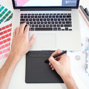 Designer using a digital drawing tablet and laptop, with colour swatches and markers beside them